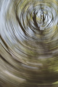 A swirling motion blur effect with circular patterns, resembling a spiral or vortex. The colors are predominantly a mix of green and brown, suggesting foliage or a forest-like setting.