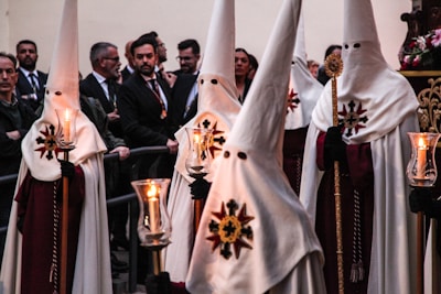A group of people dressed in traditional white garments with tall, pointed hoods and faces covered, holding candles. The attire features a prominent emblem resembling a cross with a sunburst. They are part of a procession, with onlookers present in the background.
