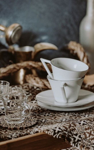 A collection of elegant white ceramic teacups neatly stacked on a saucer alongside a clear glass cup on a patterned fabric. The composition features a warm, rustic tone with hints of vintage style from the brass embellishments in the background.
