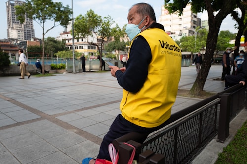 An individual wearing a bright yellow volunteer vest with '2017 Taipei Universiade' written on it is seated on a metal fence in an urban park setting. The person is wearing a blue mask and holding a tablet or phone. In the background, several people are walking or sitting, with trees and tall buildings visible under a partly cloudy sky.