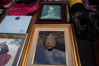A collection of framed art and objects laid out on a surface. In the center, a framed piece with a sculpted face is prominently displayed. Surrounding it are various framed artworks, including a traditional Japanese print and a green-hued painting. Nearby, a pair of black and gray hiking shoes, a bright pink piece of fabric, and a rolled-up mat in a yellow container can be seen.