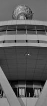 A modern architectural structure featuring geometric patterns and clean lines. The upper section appears to have a large, spherical communication dish with a lattice design. Below, the building exhibits broad, smooth panels with a series of vertical windows near the base. A security camera is mounted in an open space, facing downward.