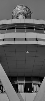 A modern architectural structure featuring geometric patterns and clean lines. The upper section appears to have a large, spherical communication dish with a lattice design. Below, the building exhibits broad, smooth panels with a series of vertical windows near the base. A security camera is mounted in an open space, facing downward.