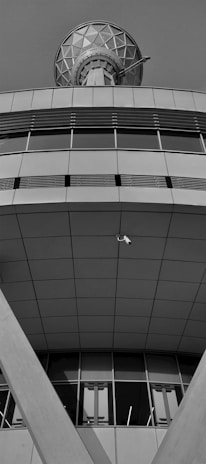 A modern architectural structure featuring geometric patterns and clean lines. The upper section appears to have a large, spherical communication dish with a lattice design. Below, the building exhibits broad, smooth panels with a series of vertical windows near the base. A security camera is mounted in an open space, facing downward.