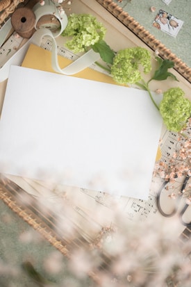 A decorative arrangement featuring a blank white sheet of paper placed over an assortment of items including sheet music, yellow envelope, scissors, and green hydrangea flowers. A white ribbon, spool, and delicate dried flowers are also present, creating a soft and artistic composition. The scene is set within a woven basket, adding texture to the image.