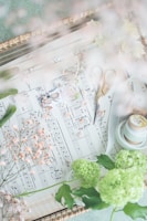 A soft, dreamy composition of sheet music adorned with delicate, small flowers. Brass scissors rest on the sheet music. Nearby are green hydrangeas and a roll of ribbon, adding to the whimsical atmosphere.