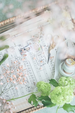 A soft, dreamy composition of sheet music adorned with delicate, small flowers. Brass scissors rest on the sheet music. Nearby are green hydrangeas and a roll of ribbon, adding to the whimsical atmosphere.
