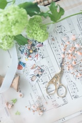 A vintage setup featuring a pair of ornate gold scissors resting on sheet music, surrounded by delicate pink flowers and green leaves. Small botanical-themed stamps add a touch of color and artistry. The overall composition is soft and artistic, with a focus on light and texture.
