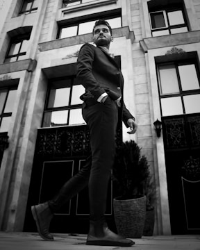 A man dressed in a tailored suit is confidently walking past an ornate building facade. The architecture features tall windows and intricate metalwork. The perspective is from below, emphasizing the man's stride and presence.