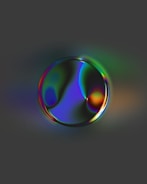 A reflective bubble-like object with an iridescent surface displaying a spectrum of colors, including shades of green, blue, and purple, against a dark gray background.