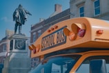 A bright yellow 74-seater coach parked outside a historic museum with excited students boarding.