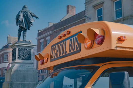 A yellow school bus is parked in front of an old building with a historical statue. The bus is prominently displayed, showing its front and distinctive orange warning lights. The background includes classic architecture with red brick elements and large windows.