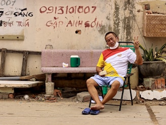 A man sits on a foldable chair next to a faded pink stone bench against a dilapidated wall. He is making a peace sign with one hand and holding food in the other. The wall behind him has hand-painted text in red and black. The ground is littered with rubble and debris. A green cooler and a packet of food sit on the bench.