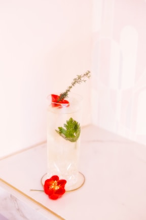A tall glass with a decorative drink featuring sprigs of rosemary and parsley on a marble surface. A red flower is placed next to the glass, adding a vibrant pop of color to the composition, which is set against a soft pastel background.