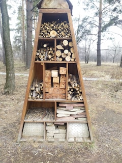 A triangular wooden structure is filled with various natural materials sorted into compartments. The materials include logs, twigs, drilled wood blocks, and bricks. The structure is set outdoors among trees, likely designed to serve as a habitat for insects.
