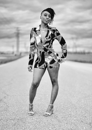 A person standing confidently on a road, wearing a patterned outfit and high heels. The background shows a cloudy sky and distant power lines, suggesting an open, rural setting. The image is in black and white.