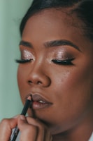 A person is having their makeup done, focusing on their face with eyes closed. They have glittery eye makeup and long eyelashes. A hand is applying lip liner to the lips, indicating a makeup session.