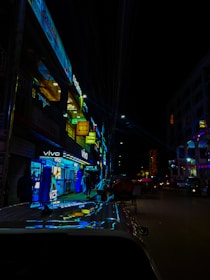 A bustling city street illuminated at night with various brightly lit shop signs, including brands like Vivo and Realme. People are seen walking along the sidewalk, and several vehicles, including rickshaws and cars, are present on the street.