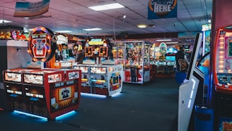A vibrant arcade filled with various game machines, including pinball, claw machines, and video games. The lighting is bright with colorful neon signs and LED lights illuminating the area. Plush toys and other prizes are visible in some machines.