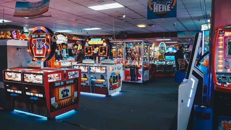 A vibrant arcade filled with various game machines, including pinball, claw machines, and video games. The lighting is bright with colorful neon signs and LED lights illuminating the area. Plush toys and other prizes are visible in some machines.