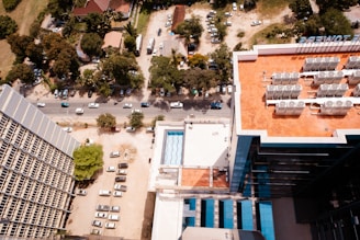 Aerial view of an urban area featuring a large building with a rooftop containing numerous air conditioning units. Surrounding the building are various trees and small structures. A road runs through the middle of the image with cars lined up along the street.
