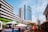 A busy urban street scene features modern high-rise buildings with blue and green glass facades. Buses and vehicles are parked along the street, with people walking nearby. An umbrella-covered stall is situated on the sidewalk, and numerous power lines stretch across the sky.