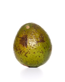 A ripe green avocado is standing upright against a white background. The surface is bumpy with noticeable brown spots and an indented top where the stem was attached.