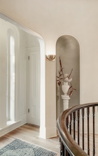 A softly lit, elegant interior corner featuring a white, curved staircase railing and a pedestal with a stylish white vase containing dried flowers. Light streaming through tall, arched windows highlights the warm wooden floor and a detailed blue and white carpet. A minimalist wall sconce adds ambiance to the neutral-toned walls.