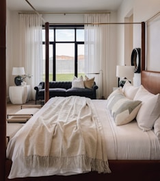 A well-decorated bedroom with a large, neatly-made bed featuring a brown leather headboard and white bedding. A soft, beige throw blanket is draped over the bed. Two white pillows and a grey accent pillow are neatly arranged. A dark blue tufted sofa sits beneath a large window with sheer white curtains. Two white side tables with lamps are positioned on either side of the bed. A round decorative mirror hangs on the wall above a lamp.