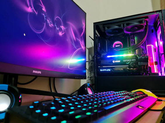 A gaming setup featuring a desktop computer tower with colorful RGB lighting, a Philips monitor displaying a vivid abstract design, a mechanical keyboard with RGB backlighting, and a pair of nearby speakers. The computer tower showcases components like an MSI GeForce RTX graphics card and NZXT cooler.
