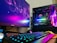 A gaming setup featuring a desktop computer tower with colorful RGB lighting, a Philips monitor displaying a vivid abstract design, a mechanical keyboard with RGB backlighting, and a pair of nearby speakers. The computer tower showcases components like an MSI GeForce RTX graphics card and NZXT cooler.
