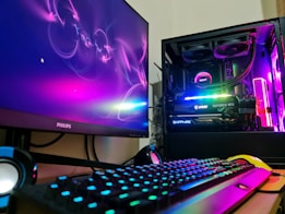 A gaming setup featuring a desktop computer tower with colorful RGB lighting, a Philips monitor displaying a vivid abstract design, a mechanical keyboard with RGB backlighting, and a pair of nearby speakers. The computer tower showcases components like an MSI GeForce RTX graphics card and NZXT cooler.