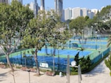 A sports complex featuring multiple tennis courts surrounded by trees and urban buildings in the background. The courts are enclosed by fencing and covered with blue and green surfaces. Benches and a clock are visible near the courts, with tall buildings rising in the distance.