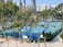 A sports complex featuring multiple tennis courts surrounded by trees and urban buildings in the background. The courts are enclosed by fencing and covered with blue and green surfaces. Benches and a clock are visible near the courts, with tall buildings rising in the distance.