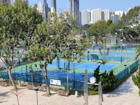 A sports complex featuring multiple tennis courts surrounded by trees and urban buildings in the background. The courts are enclosed by fencing and covered with blue and green surfaces. Benches and a clock are visible near the courts, with tall buildings rising in the distance.