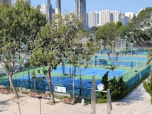 A sports complex featuring multiple tennis courts surrounded by trees and urban buildings in the background. The courts are enclosed by fencing and covered with blue and green surfaces. Benches and a clock are visible near the courts, with tall buildings rising in the distance.