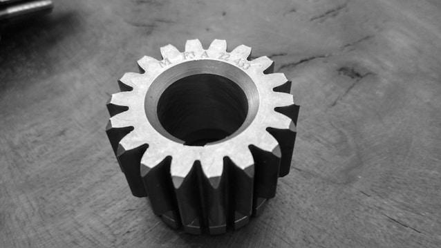A monochrome image of a metallic gear resting on a textured surface. The gear has sharp, evenly spaced teeth with an engraved marking on the top face.