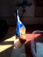 A brown egg is placed on a countertop, partially illuminated by sunlight streaming through a window. Beside the egg, a sealed bag of flour stands upright, casting a shadow. The countertop has a wooden cutting board and a red cloth. A plastic container is partially visible in the foreground.