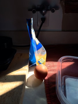 A brown egg is placed on a countertop, partially illuminated by sunlight streaming through a window. Beside the egg, a sealed bag of flour stands upright, casting a shadow. The countertop has a wooden cutting board and a red cloth. A plastic container is partially visible in the foreground.