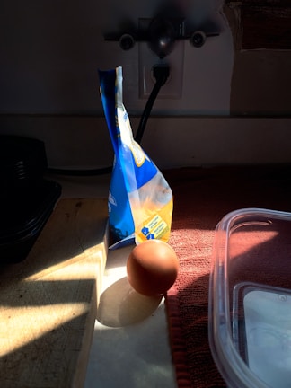 A brown egg is placed on a countertop, partially illuminated by sunlight streaming through a window. Beside the egg, a sealed bag of flour stands upright, casting a shadow. The countertop has a wooden cutting board and a red cloth. A plastic container is partially visible in the foreground.