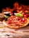 A rustic pepperoni pizza is placed on a wooden surface, topped with red chillies and a mix of vegetables. In the background, there are fresh tomatoes, a purple onion, and bowls filled with ingredients such as peppercorns and cheese. Flames create a warm, fiery backdrop.
