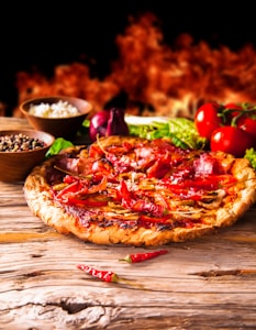 A rustic pepperoni pizza is placed on a wooden surface, topped with red chillies and a mix of vegetables. In the background, there are fresh tomatoes, a purple onion, and bowls filled with ingredients such as peppercorns and cheese. Flames create a warm, fiery backdrop.