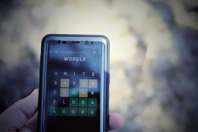 A close-up view of a smartphone displaying the Wordle game interface, with a hand holding the phone. The screen shows words arranged in a grid with various letters highlighted in green and yellow.