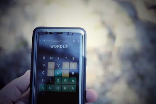 A close-up view of a smartphone displaying the Wordle game interface, with a hand holding the phone. The screen shows words arranged in a grid with various letters highlighted in green and yellow.