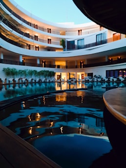 A multi-storied building with an open courtyard featuring a swimming pool in the center. The design is modern, with circular balconies and sleek lines. The pool reflects the warm lights from the windows and balconies, creating a serene and luxurious ambiance. Lush greenery accents the space along the lower level.