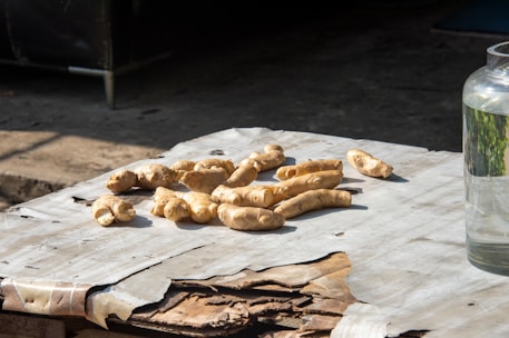 Several pieces of fresh ginger root are placed on a table covered with worn paper. The setting is outdoors, indicated by the shadow and sunlight. To the right, a glass jar is partially visible, filled with clear liquid.