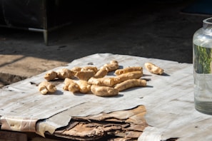 Several pieces of fresh ginger root are placed on a table covered with worn paper. The setting is outdoors, indicated by the shadow and sunlight. To the right, a glass jar is partially visible, filled with clear liquid.