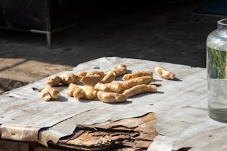 Several pieces of fresh ginger root are placed on a table covered with worn paper. The setting is outdoors, indicated by the shadow and sunlight. To the right, a glass jar is partially visible, filled with clear liquid.
