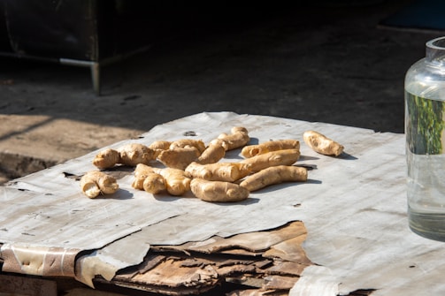 Several pieces of fresh ginger root are placed on a table covered with worn paper. The setting is outdoors, indicated by the shadow and sunlight. To the right, a glass jar is partially visible, filled with clear liquid.