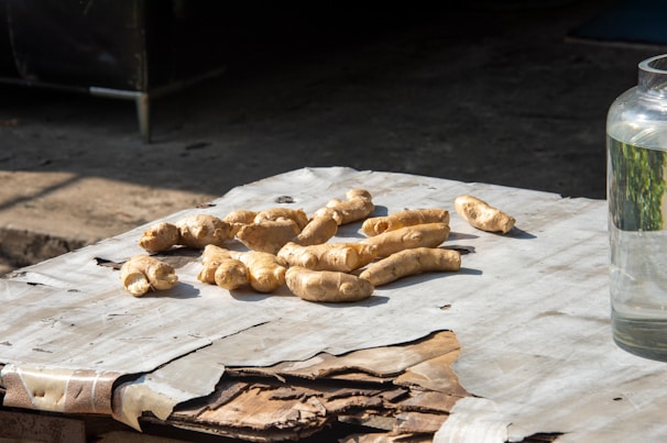 Several pieces of fresh ginger root are placed on a table covered with worn paper. The setting is outdoors, indicated by the shadow and sunlight. To the right, a glass jar is partially visible, filled with clear liquid.